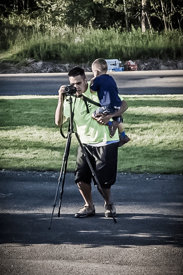 Jason holding a child while looking through a camera on a tripod.