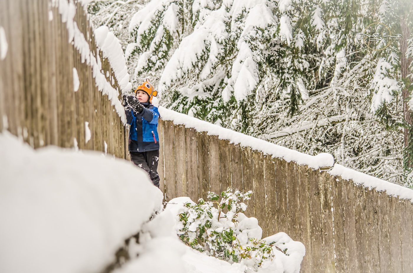 Child in Fish Hat Reaching for Snow on Wooden Fence